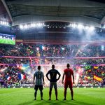 Messi, Mbappé, and Kane stand on a pitch facing a packed stadium of fans waving international flags